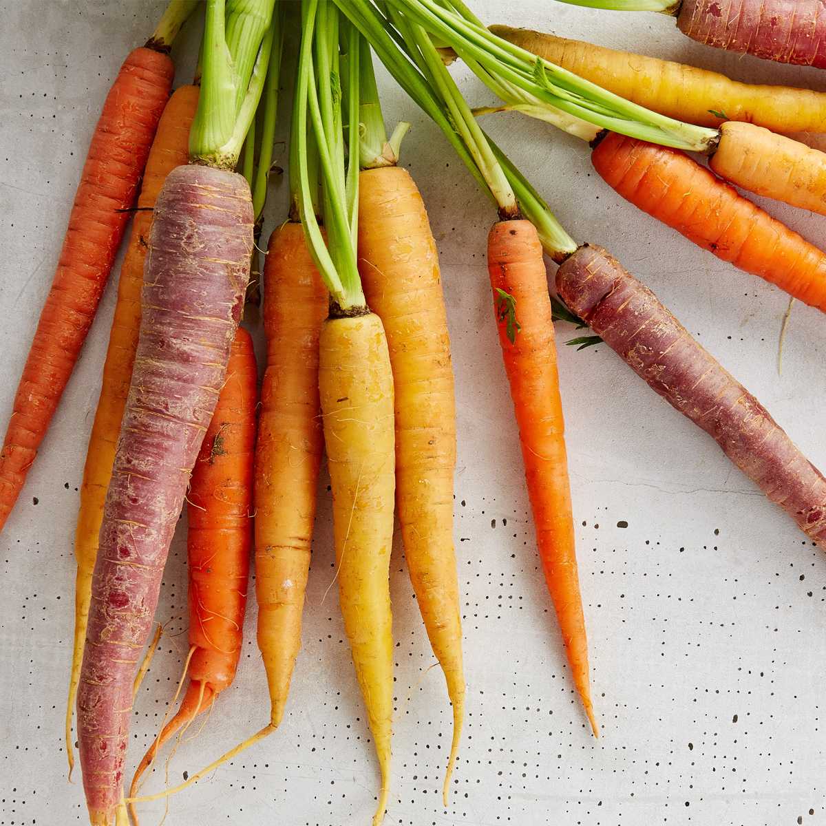 Overhead view of colorful rainbow carrots with green stems on a light speckled surface, vibrant orange purple and yellow